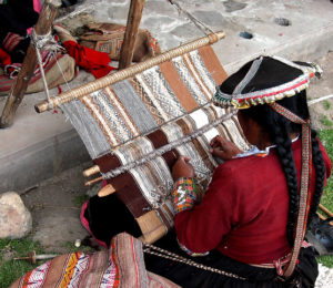 Peruvian woman dressed in traditional clothing, sitting on the ground, weaving a scaffold piece with a backstrap loom